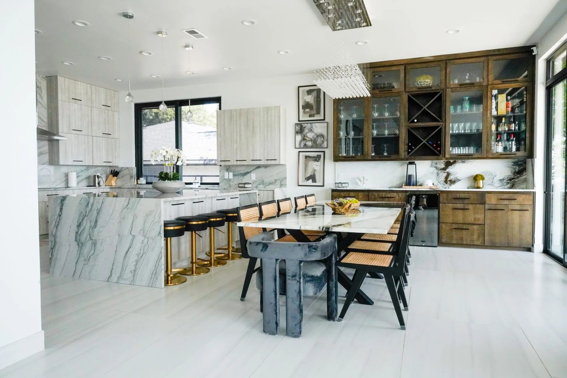 Modern kitchen with a large marble island, wooden cabinets, and a dining table. Black and gold bar stools and chairs are present.