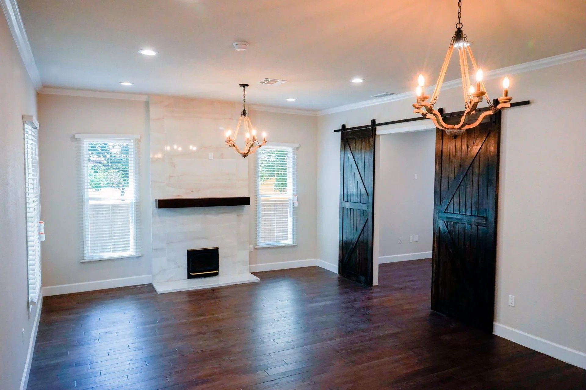 An empty living room with dark wood floors, a white fireplace, two chandeliers, and barn door-style doors.
