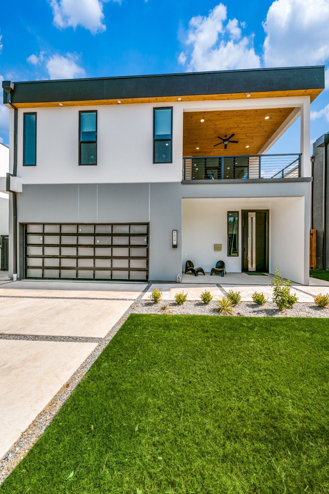 Modern two-story house with white and gray exterior, balcony, glass garage door, and green lawn. Concrete driveway and small shrubs in front.