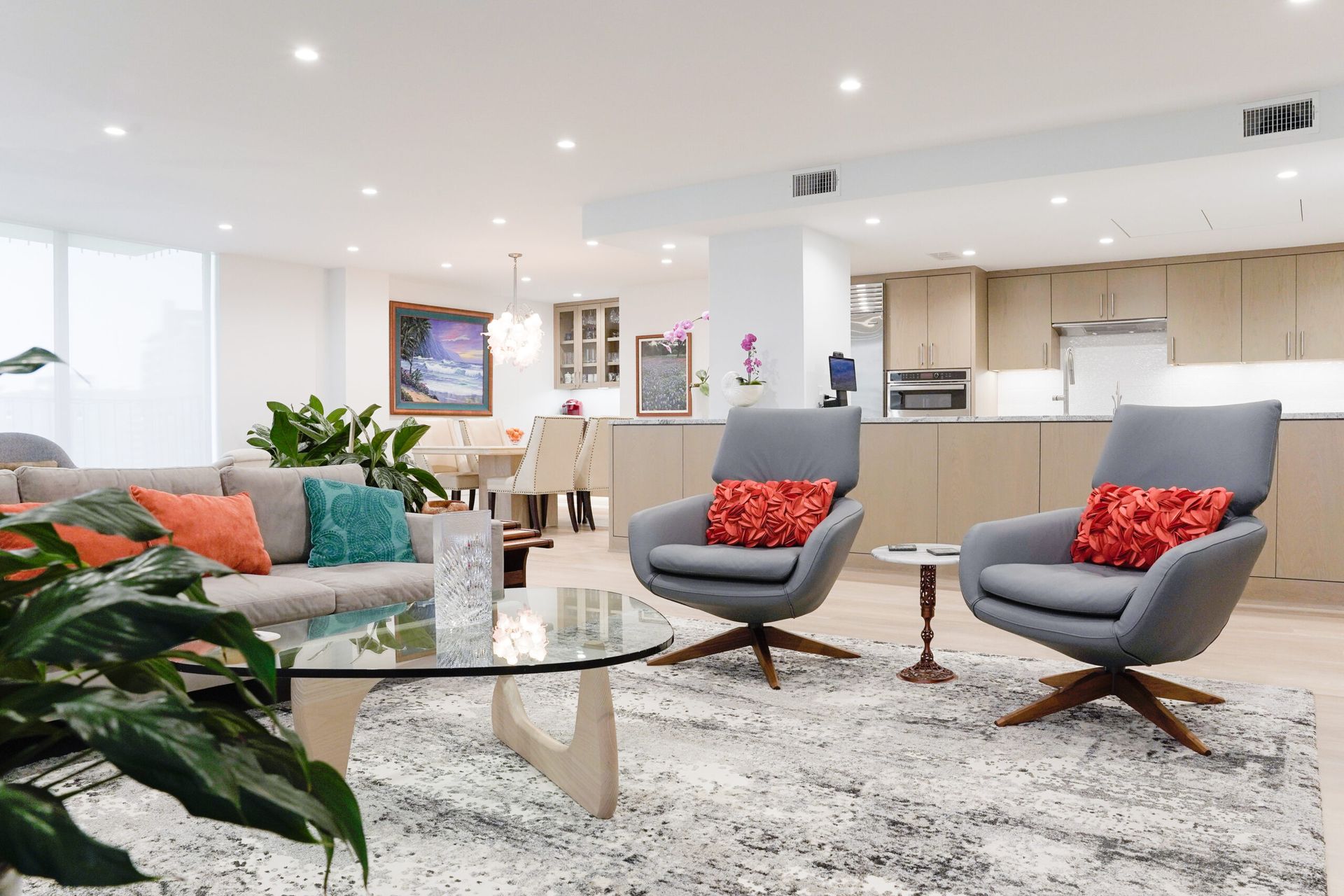 Modern living room with open kitchen. Gray chairs with red pillows face a glass coffee table; a light-colored sofa has teal and orange pillows.