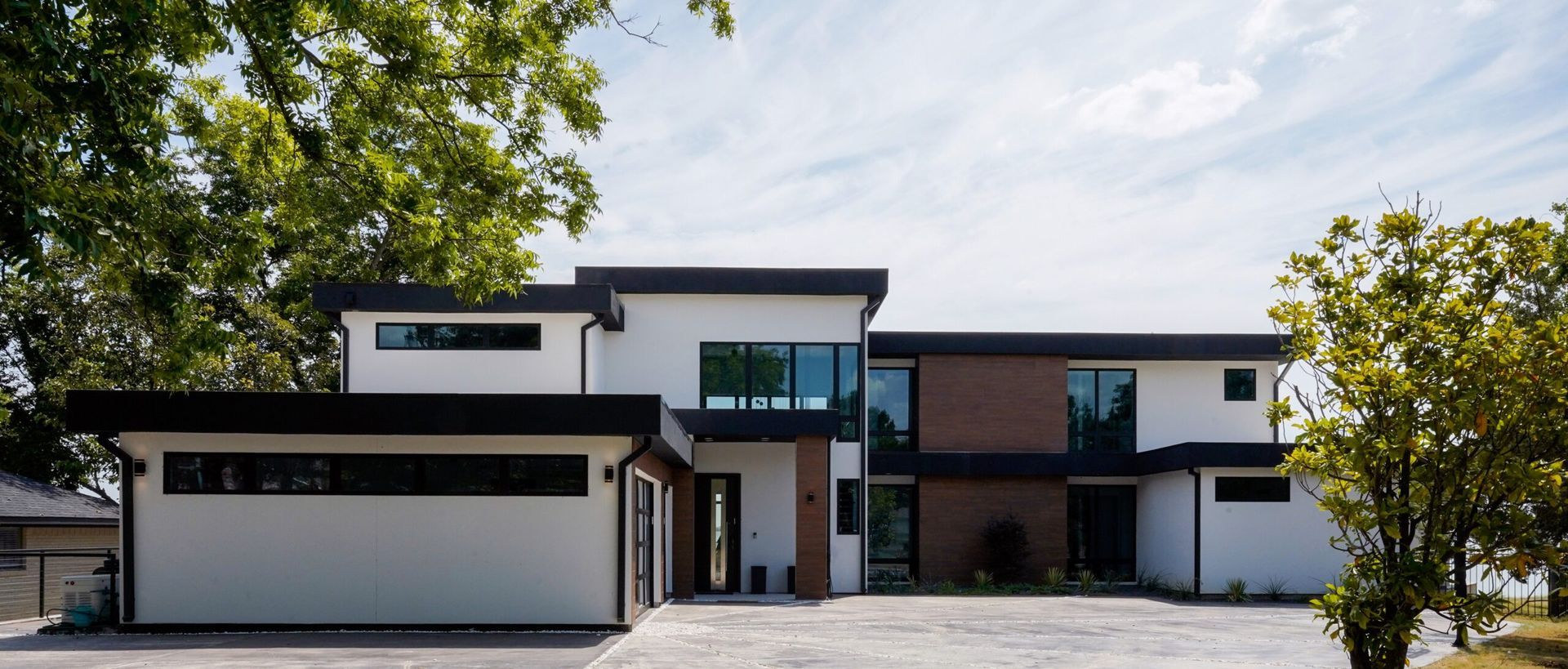 Modern white house with black trim, windows, and accents.  A two-car garage and trees frame the home on a sunny day.