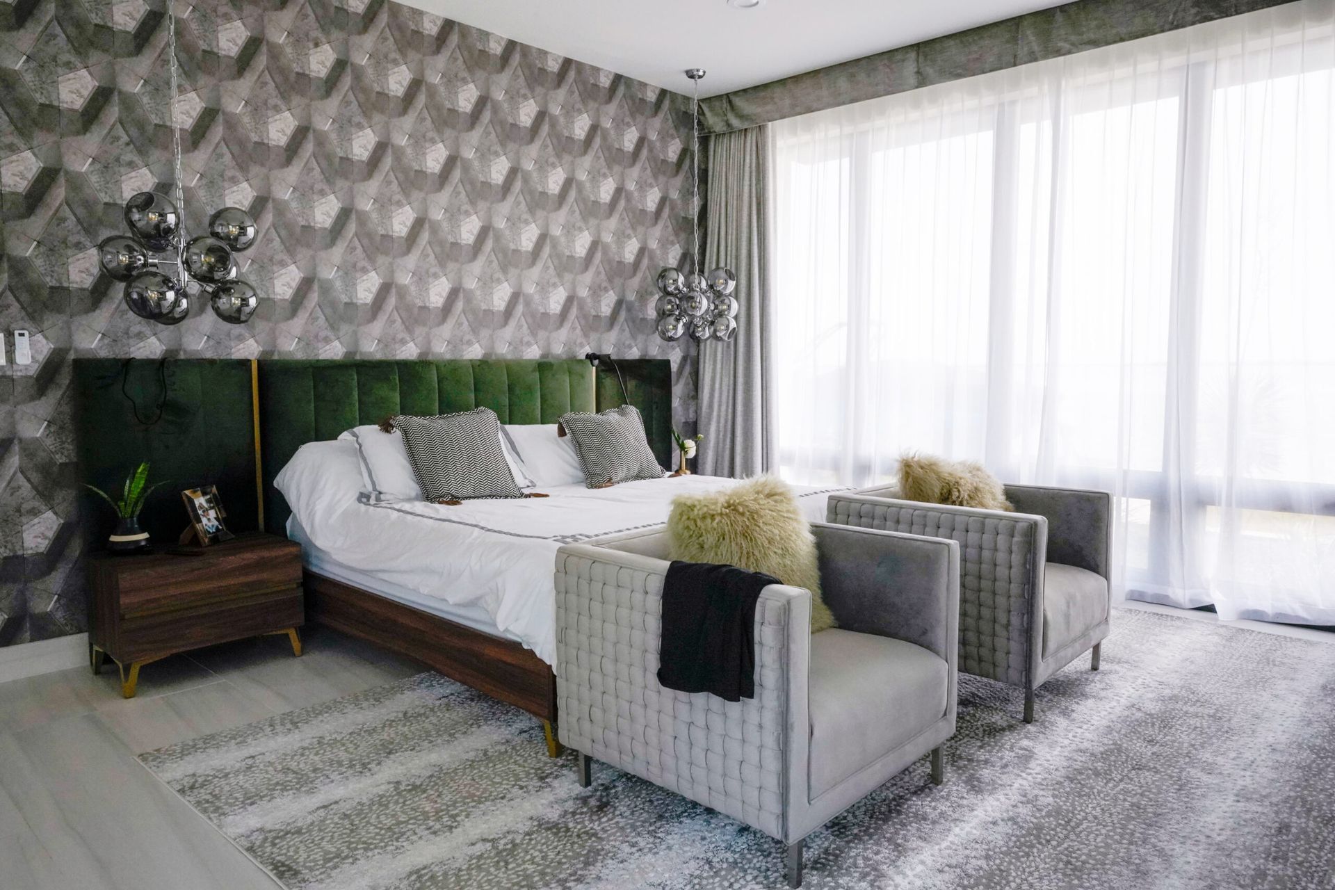 A modern bedroom with a large bed, velvet green headboard, two gray chairs, and a geometric patterned wallpaper.
