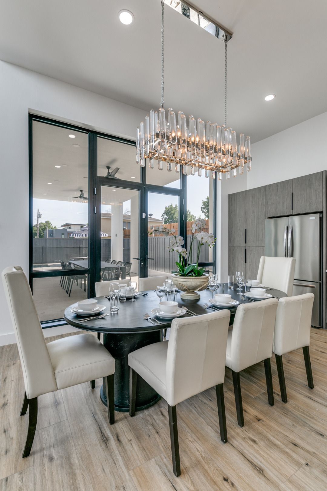 Elegant dining room with a large oval table set for dinner, beneath a crystal chandelier. Large windows reveal an outdoor patio.