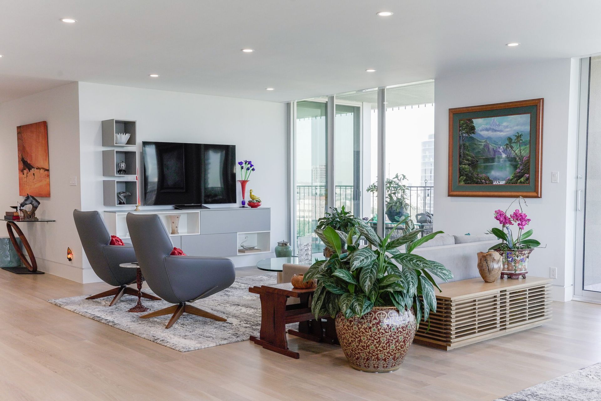 Modern living room with grey armchairs, a TV, and decorative plants. Light wood flooring, white walls, and large windows with a city view.