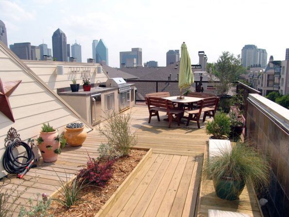 Rooftop deck with cityscape view, featuring a dining area, grill, and planters with various plants. Wooden deck with a tan and beige color scheme.
