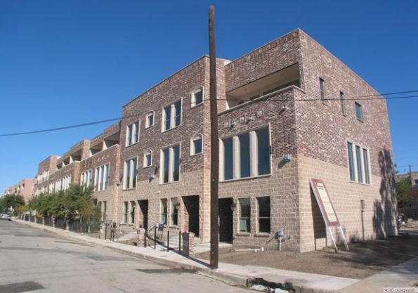 Row of multi-story brick townhouses under a clear blue sky. The street slopes downward.