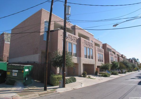 Row of brick townhomes on a sunny street. Two-car garage and multiple windows on each unit. Power lines and dumpsters in the foreground.