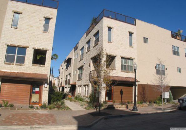 Modern three-story townhouses with tan brick exteriors and rooftop patios under a clear blue sky.