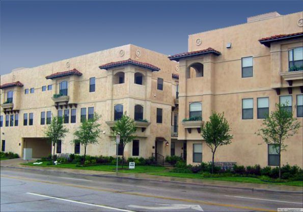 Apartment complex with light tan stucco walls and red tile roof accents, set on a street with small trees and grass.