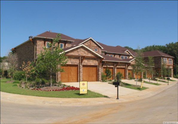Townhomes with brick exteriors and wood garage doors on a curving street under a clear blue sky.