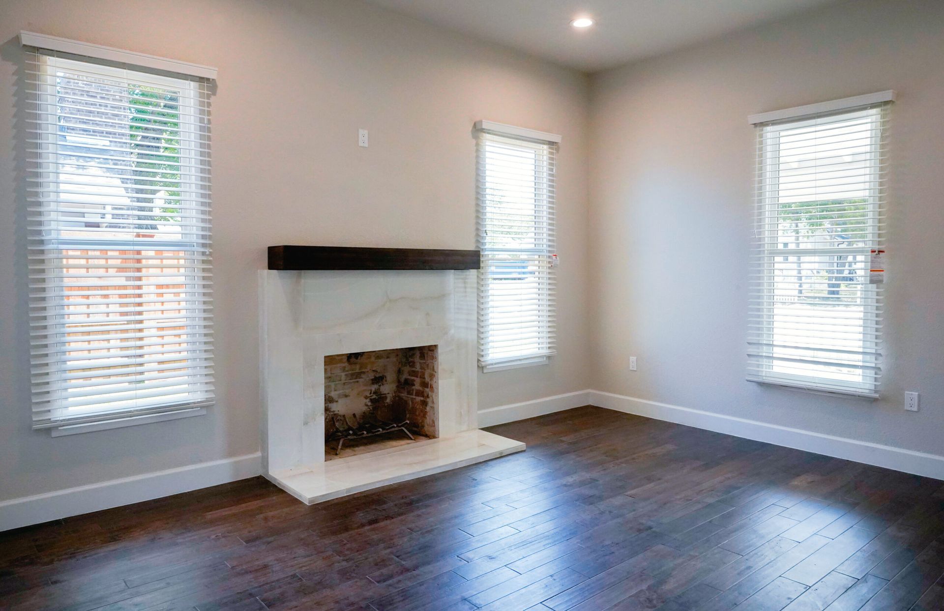 Empty room with dark wood floors, white fireplace, and three windows with blinds.