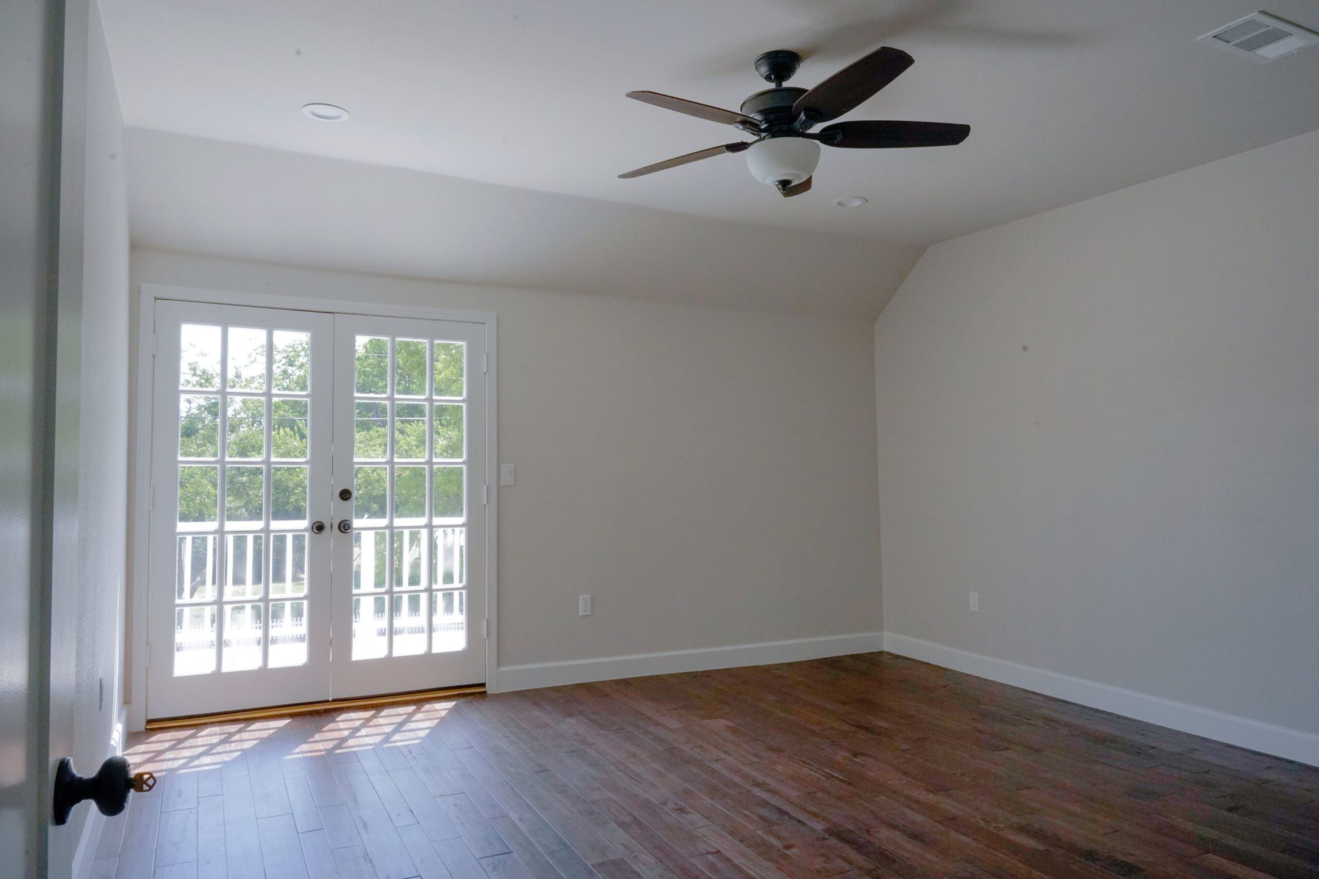 Empty room with french doors leading to a balcony. Dark wood floors, off-white walls, and a ceiling fan.