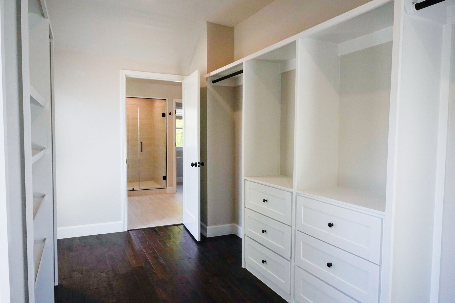 Walk-in closet with white built-in shelving and drawers. A doorway leads to a light-filled bathroom.