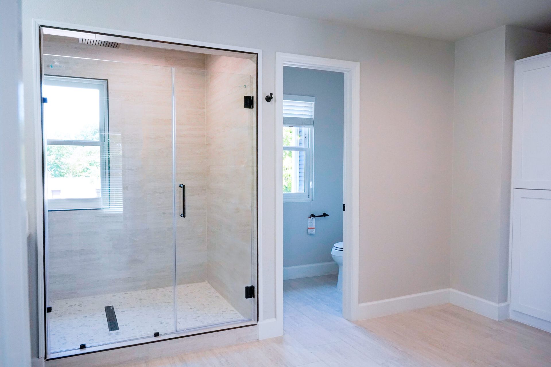 Modern bathroom with a glass shower enclosure, open doorway to a toilet, and a white cabinet. Light walls and flooring.