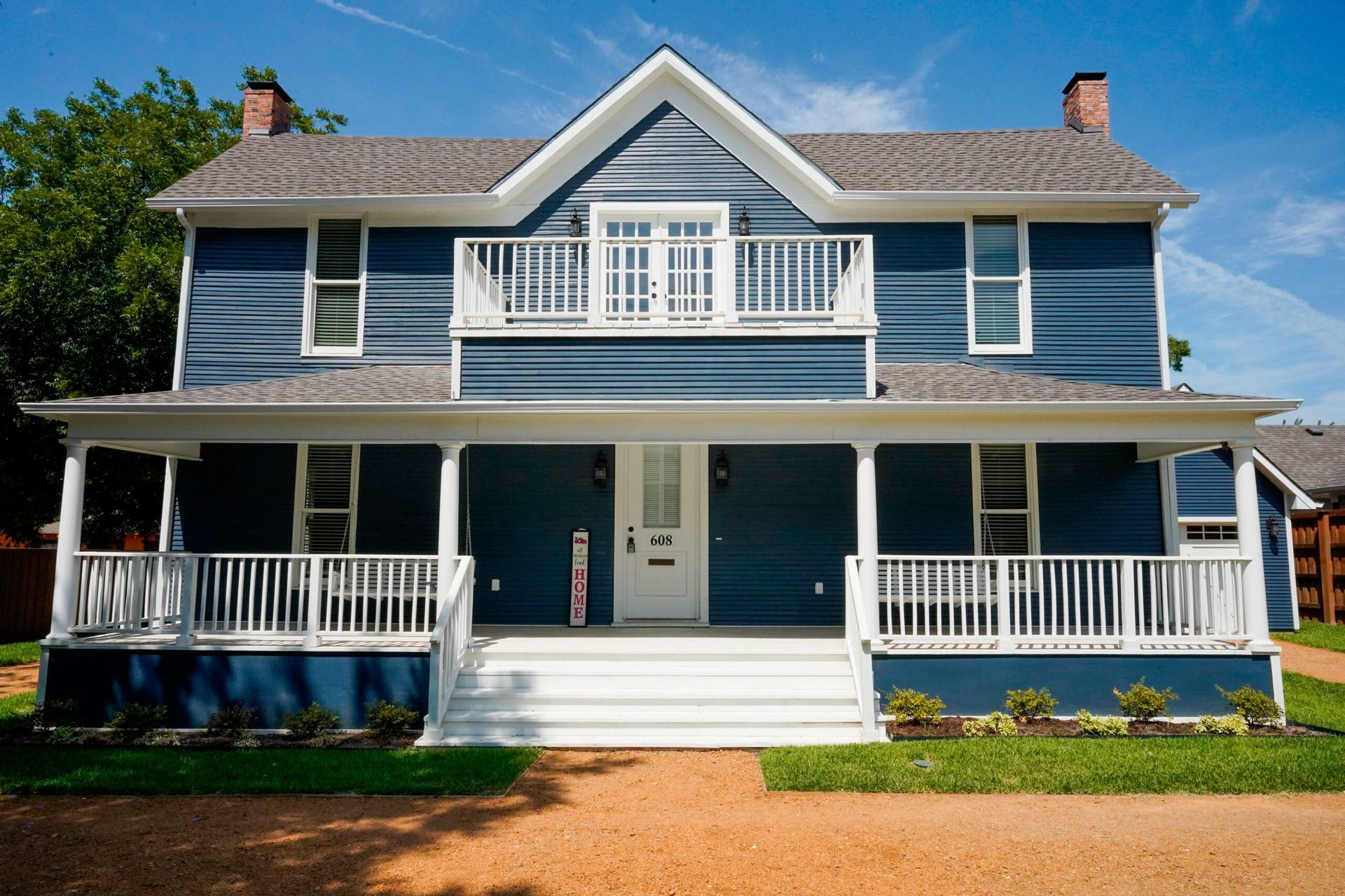 Two-story blue house with white trim, porch, and a balcony, on a sunny day.