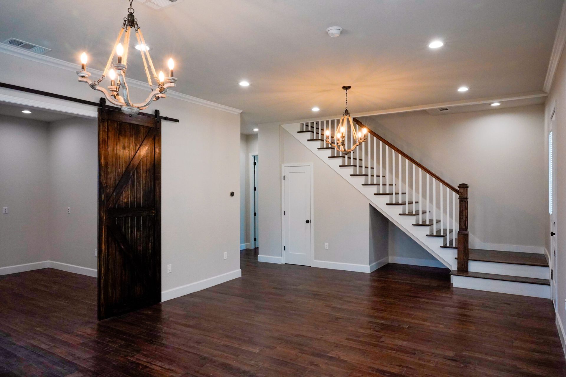 Spacious interior with dark wood floors, white walls, and a wooden barn door. A staircase and chandeliers are also visible.