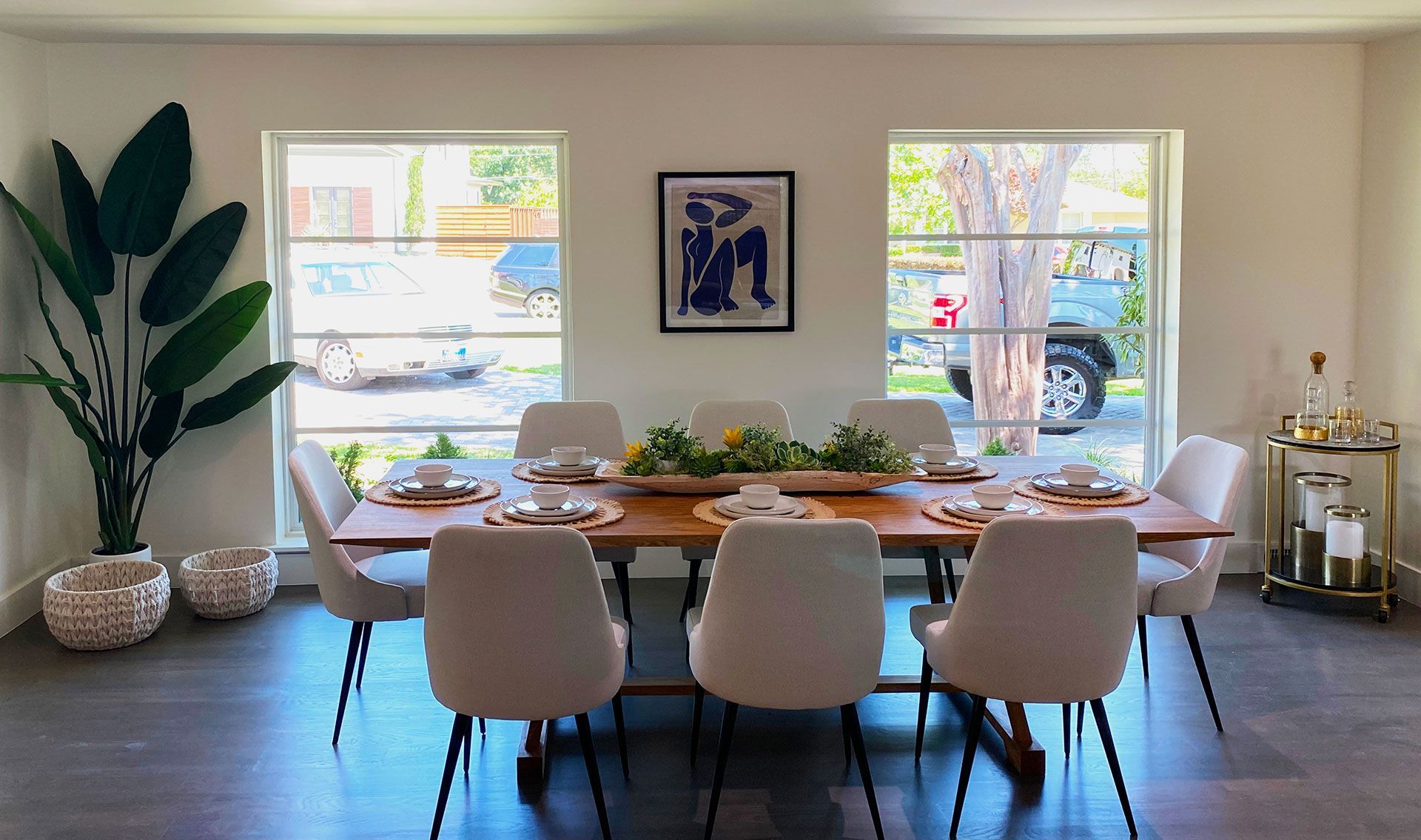 Dining room with a long wooden table set for guests, with white chairs, and large windows.