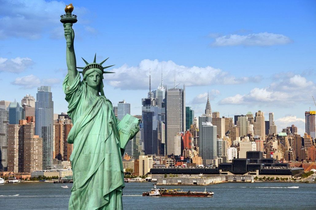 Statue of Liberty with cityscape in background, blue sky, water in foreground.