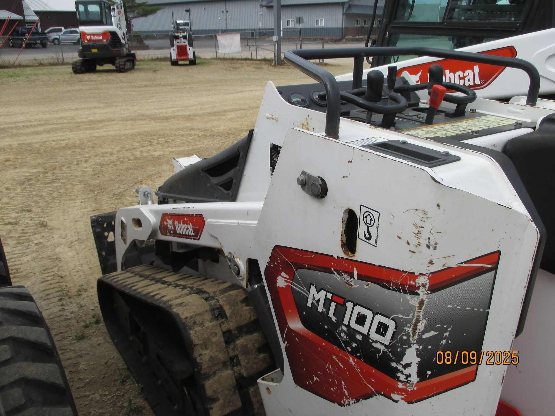 A close-up of a white and red Bobcat MT100 compact track loader on a dirt lot. The machine has tracks, controls, and the Bobcat logo.