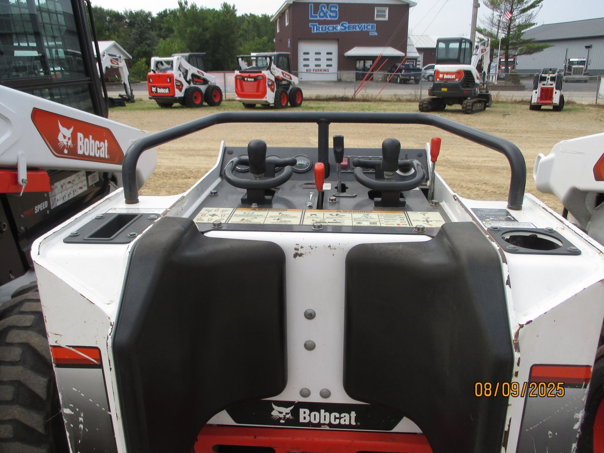 Bobcat skid-steer controls with two joysticks, a metal bar, and the Bobcat logo. Other Bobcats are visible in the background.