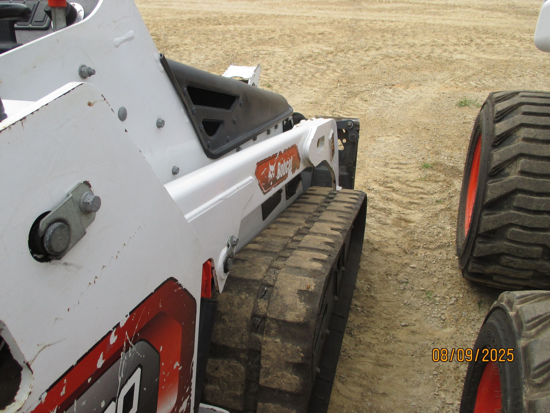 White and orange Bobcat compact track loader on a sandy surface, with a track in the foreground. The date
