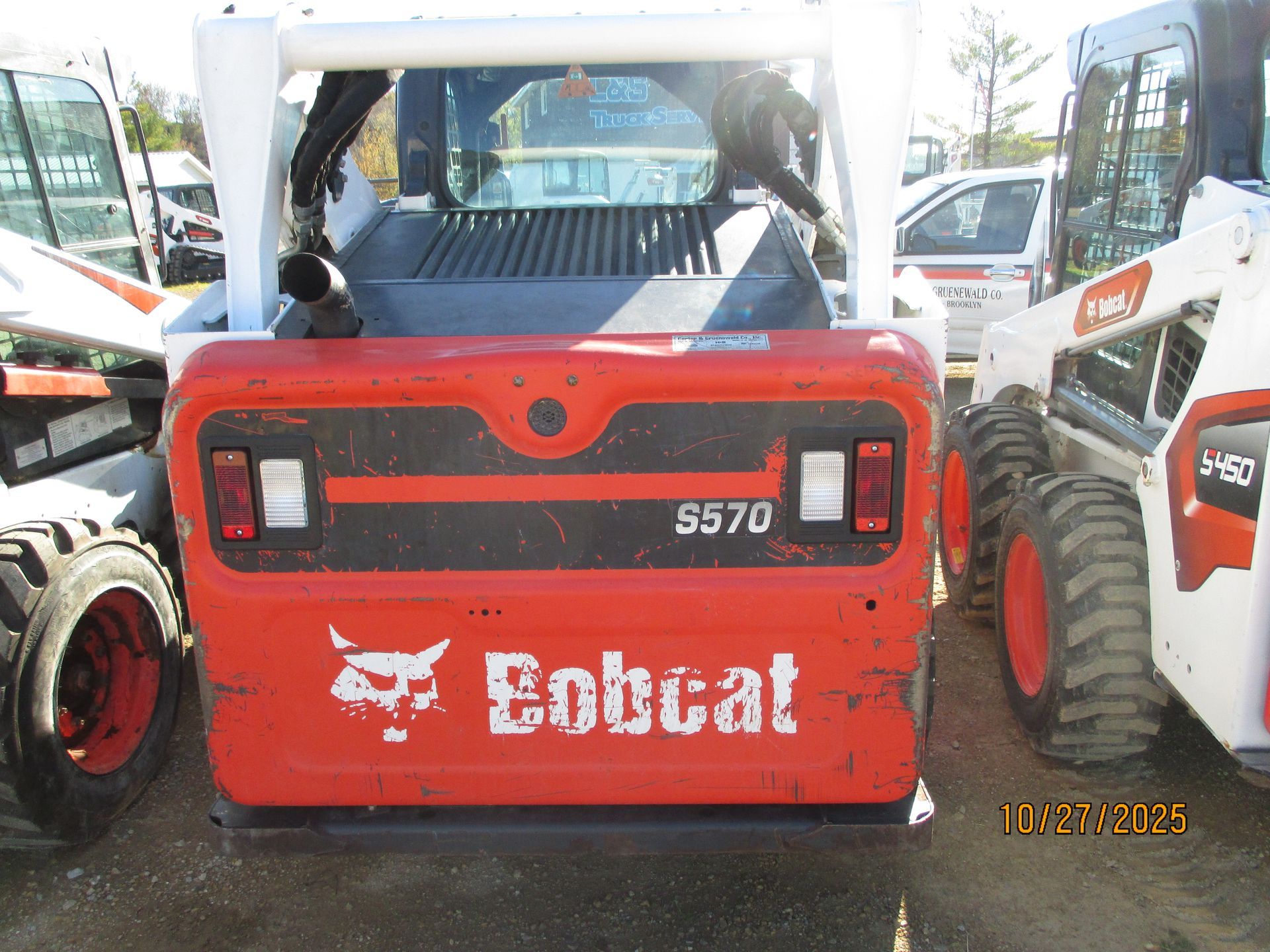 Rear view of an orange Bobcat S570 skid-steer loader with white lettering and taillights, parked outside with other machines.