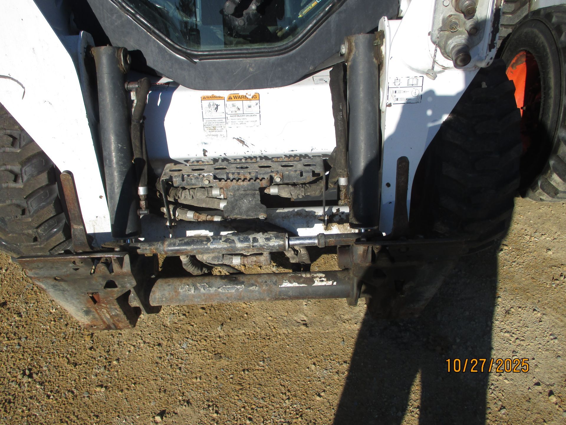 Skid-steer loader's rear view, showing tire, metal frame, and hydraulic components in a gravel setting.