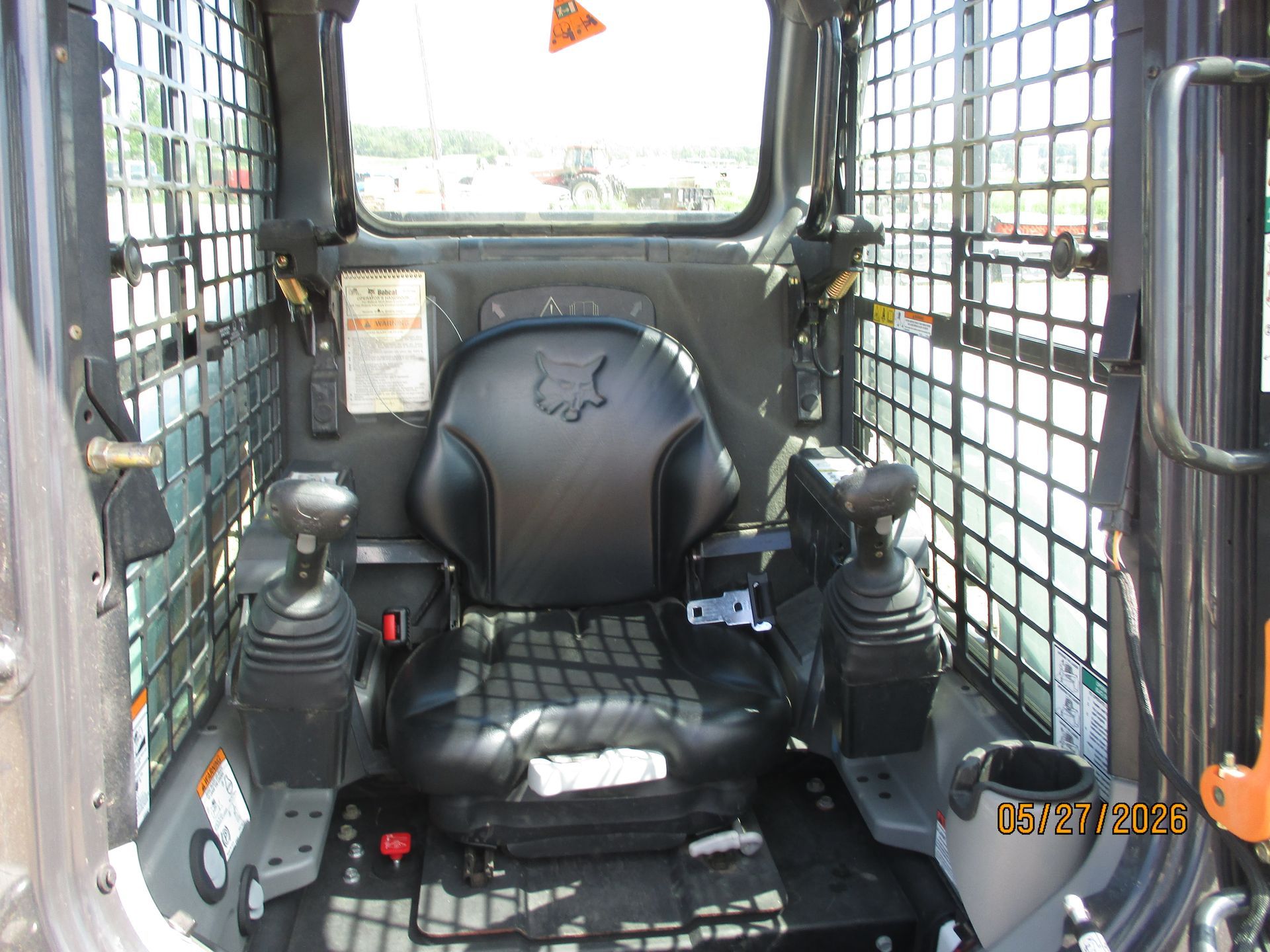 Inside a skid steer cab with black seat, control joysticks, and wire-mesh windows