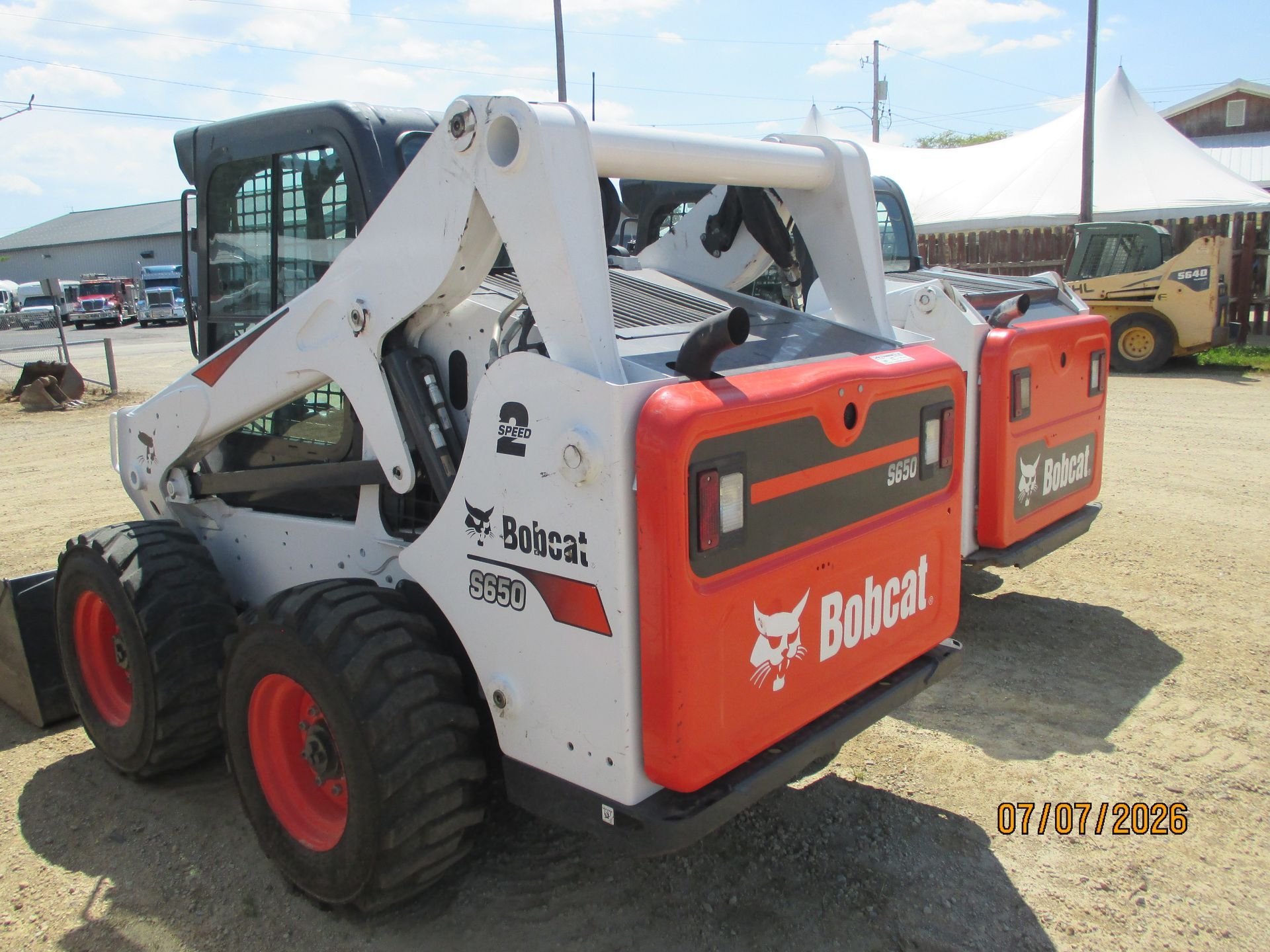 White Bobcat skid-steer loader with orange rear panel parked outdoors on gravel at an event.