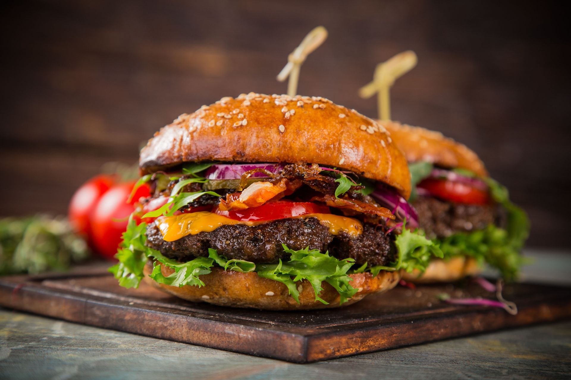 Two gourmet burgers with sesame seed buns on a wooden board, with tomatoes in the background.