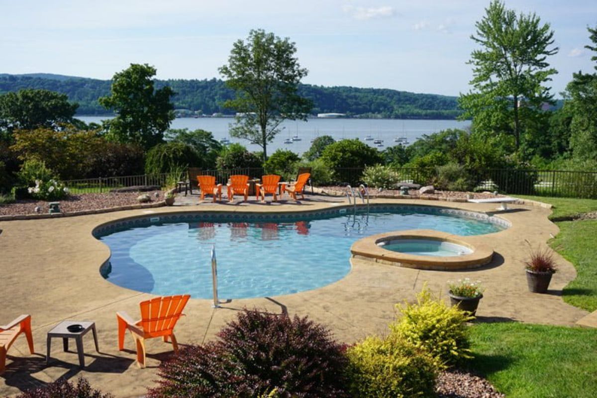 A large swimming pool surrounded by chairs and a hot tub overlooking a lake