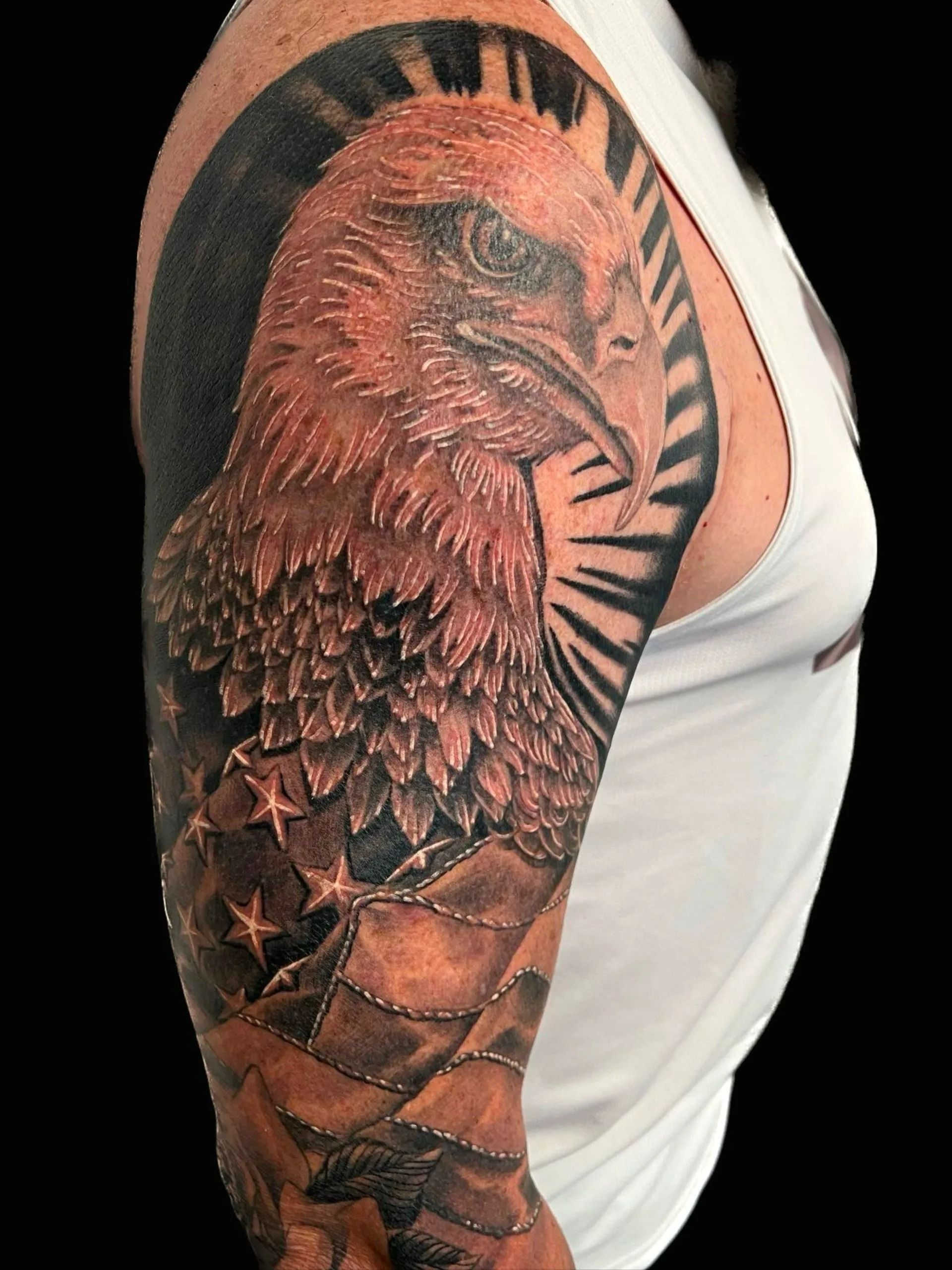 Tattoo of a bald eagle with American flag on upper arm. Black and gray, with a white tank top.