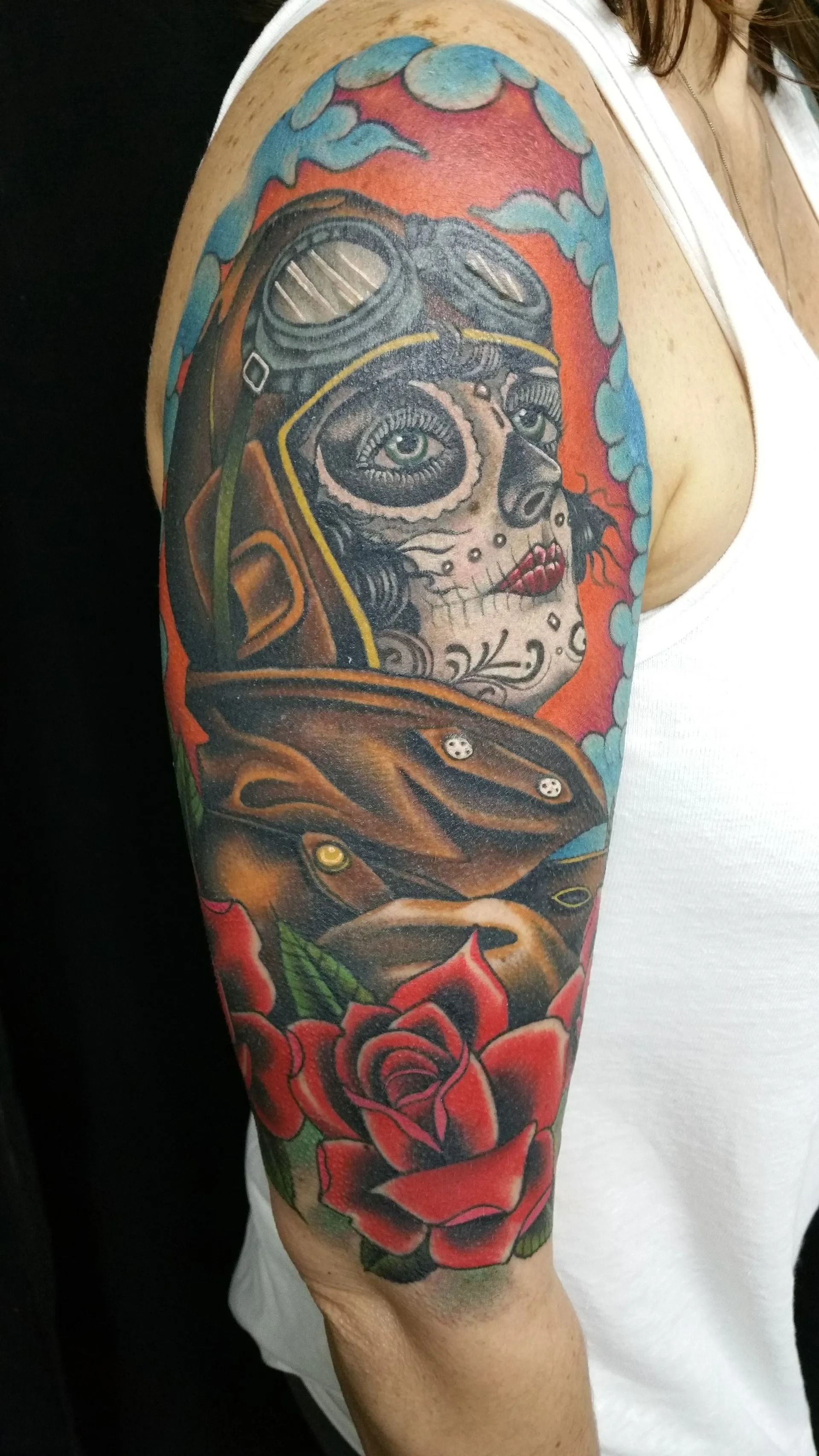 Tattoo on arm: sugar skull in pilot gear, surrounded by roses and blue/orange clouds.