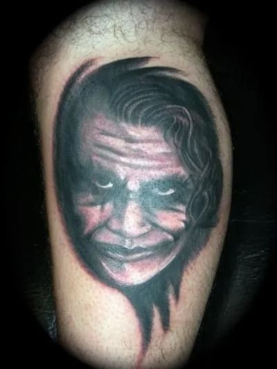 Tattoo of The Joker's face, with green and red accents, on a leg. Dark hair, sinister smile, intense gaze.