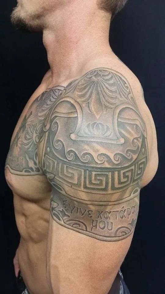 Tattooed muscular arm and shoulder with Greek-inspired design. Includes text, geometric patterns, and waves.