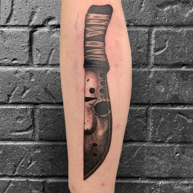 Tattoo of a knife resembling Jason Voorhees' mask, on a person's leg, against a dark brick wall.