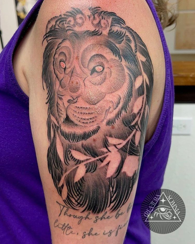 Tattoo of a lion's face with a floral wreath on a person's upper arm. Text reads 