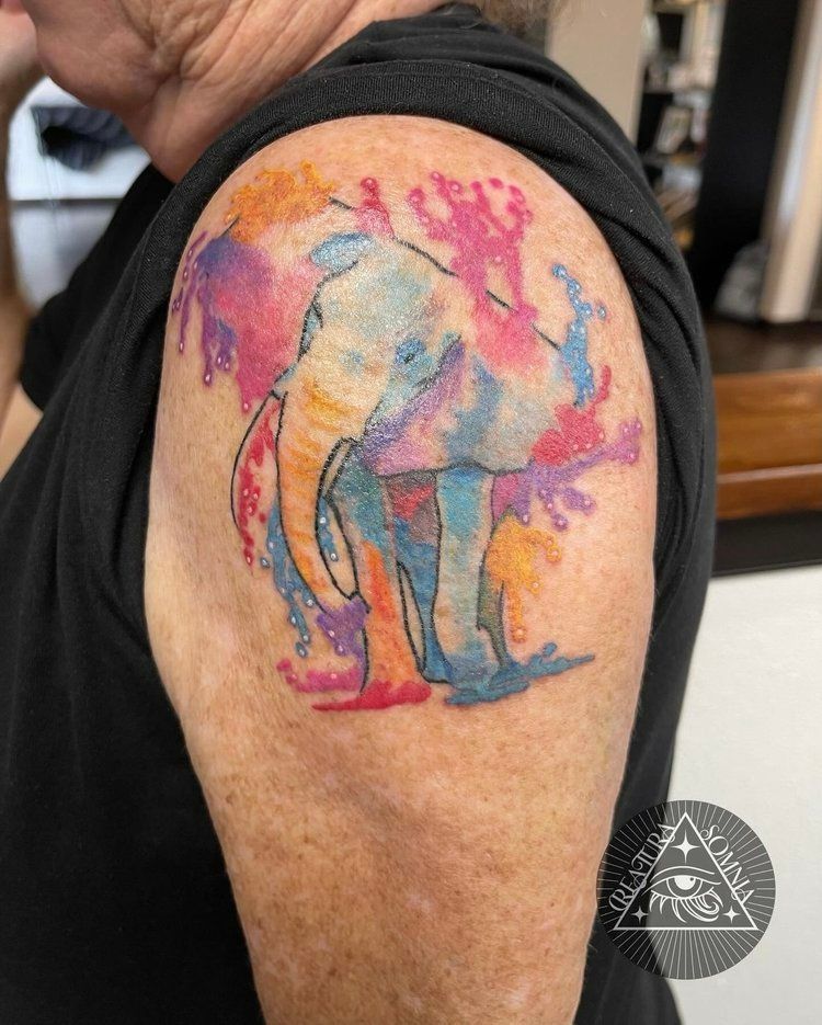 Watercolor elephant tattoo on a person's upper arm, with colorful splashes and outlines.