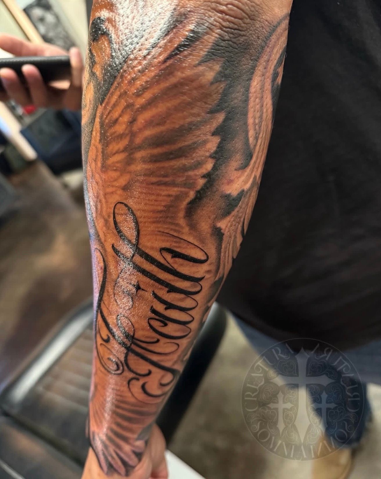 Close-up of a tattooed forearm with a black script name and black-and-brown feather design