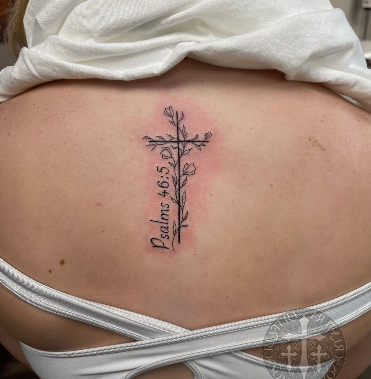Tattoo of a cross with floral design and Psalm 46:5 on a person's back.