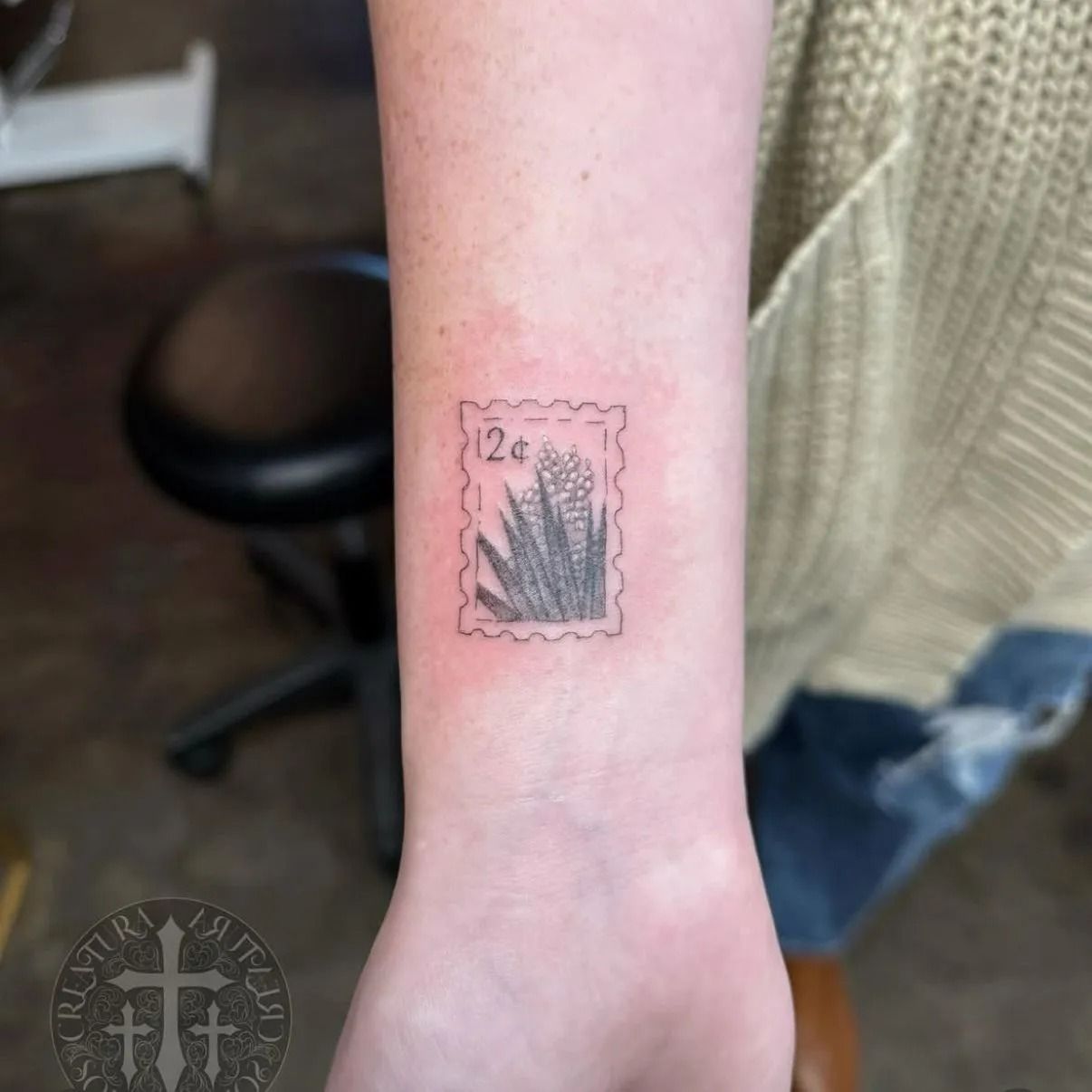Tattoo of a postage stamp with a floral design on a wrist, in black ink.