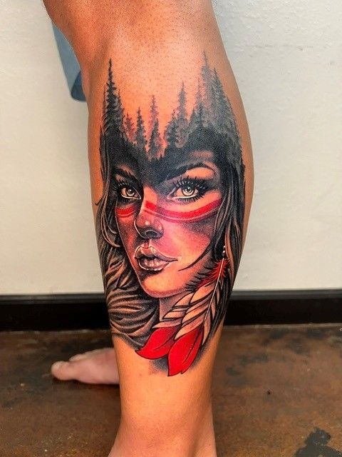 Tattoo of a woman's face with red war paint, forest silhouette above, red feathers.