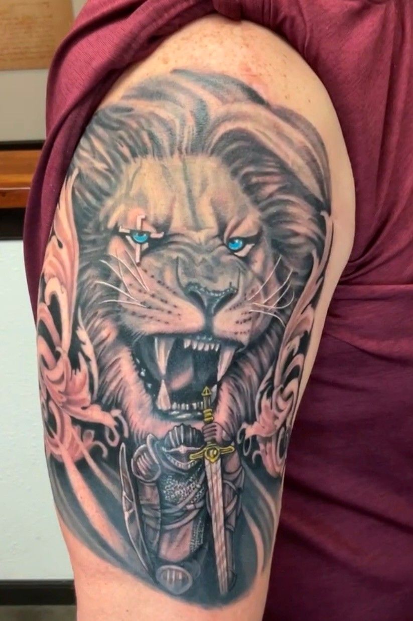 Lion tattoo with blue eyes, snarling, with a sword and armor. Arm visible, wearing burgundy shirt.