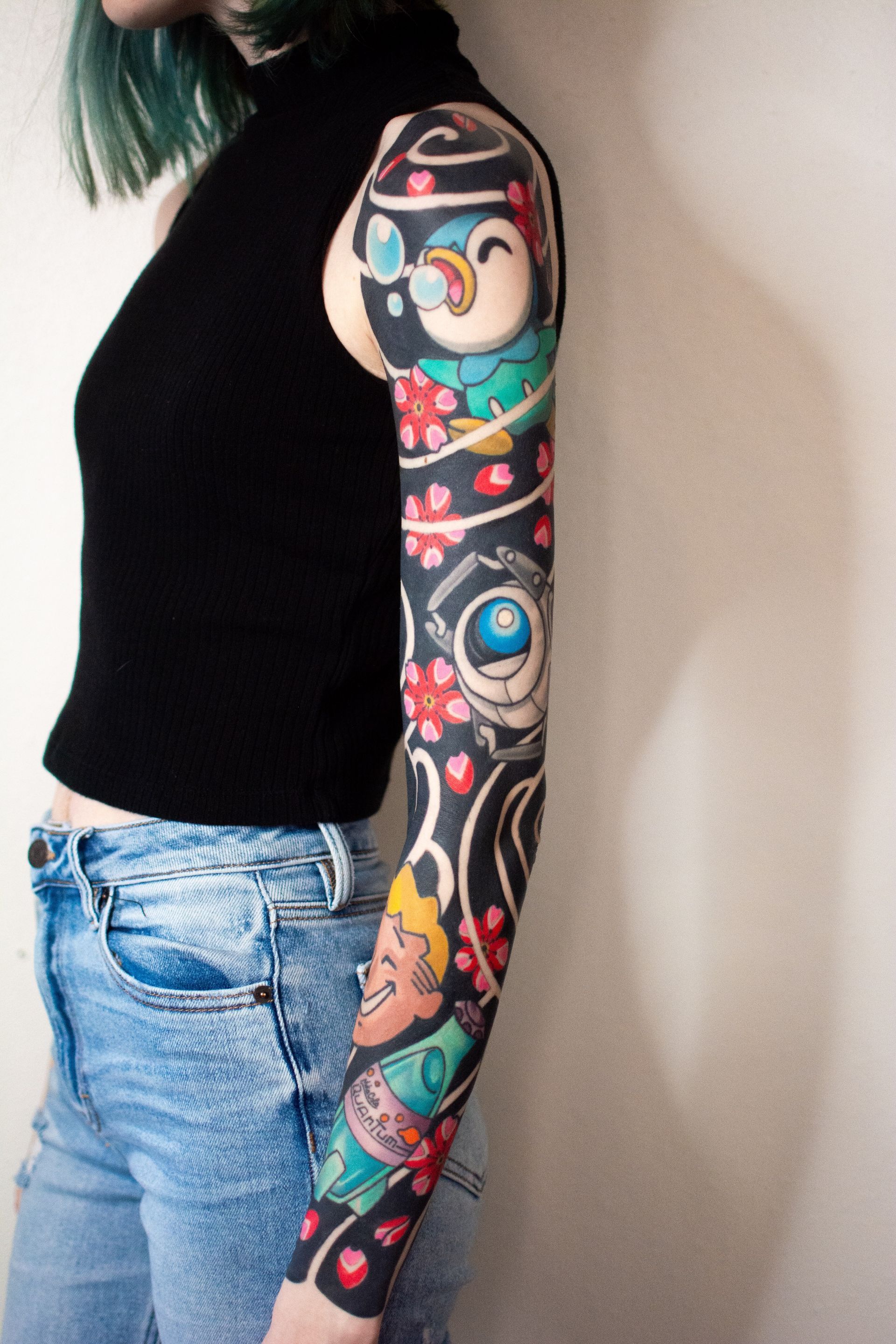 Woman with full sleeve tattoo, featuring cartoon characters and cherry blossoms. She wears a black top and jeans.