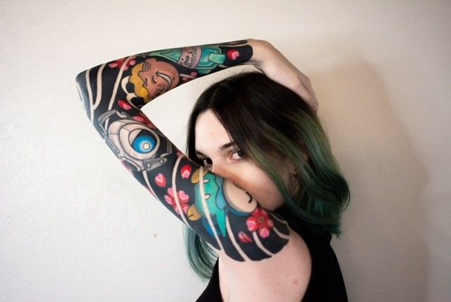 Woman with black and colorful arm tattoos poses with arm bent, against a white background.