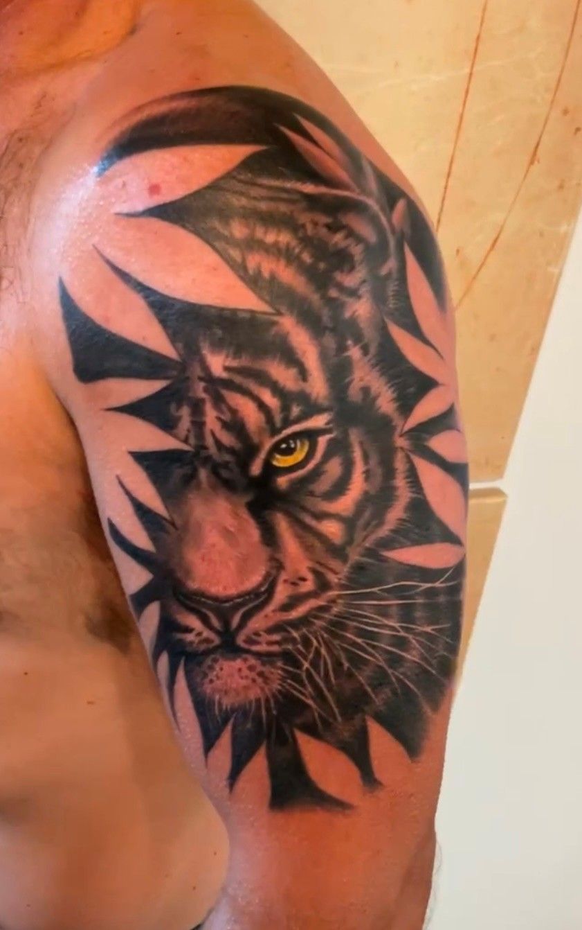 Tattoo of a tiger's face on a person's upper arm. The tiger has yellow eyes and black stripes. Black geometric shapes surround it.