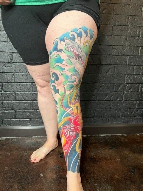 Leg tattoo: a shark emerging from blue and green waves, over a red and yellow flower.