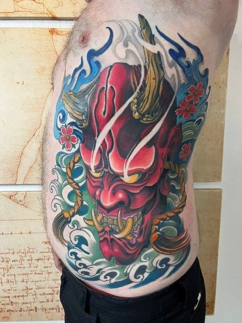 Tattoo of a red demon mask with horns and teeth, surrounded by blue water waves and flowers on a torso.