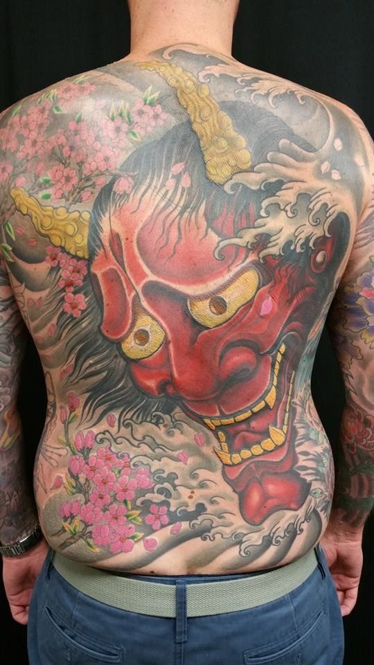 Full back tattoo: red oni mask with golden horns, surrounded by pink flowers and waves.