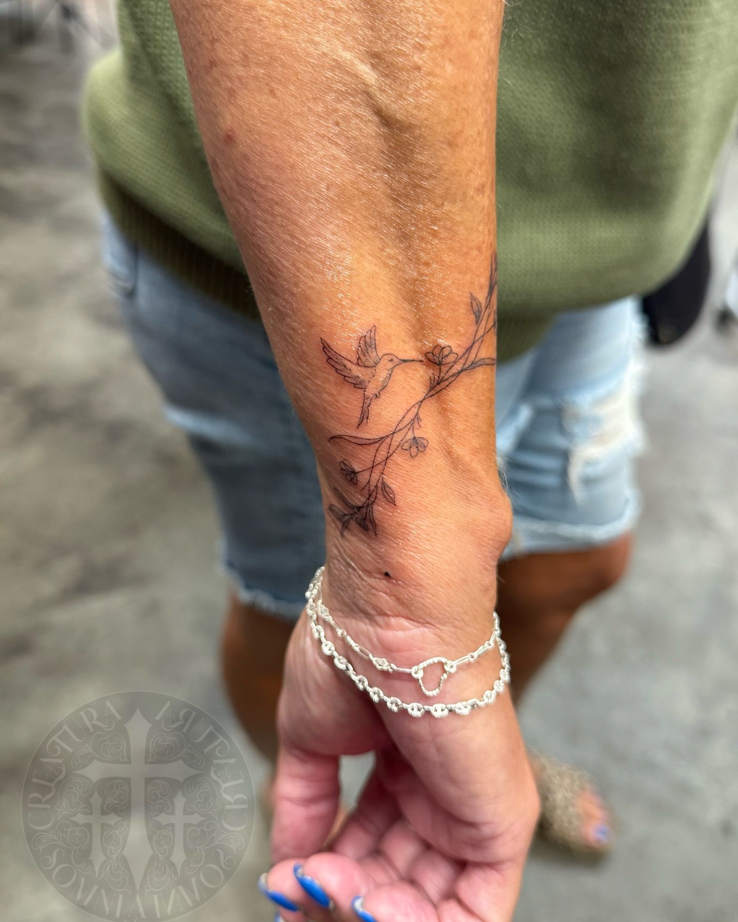 Wrist tattoo of a hummingbird branch, person wearing silver bracelet and denim shorts.